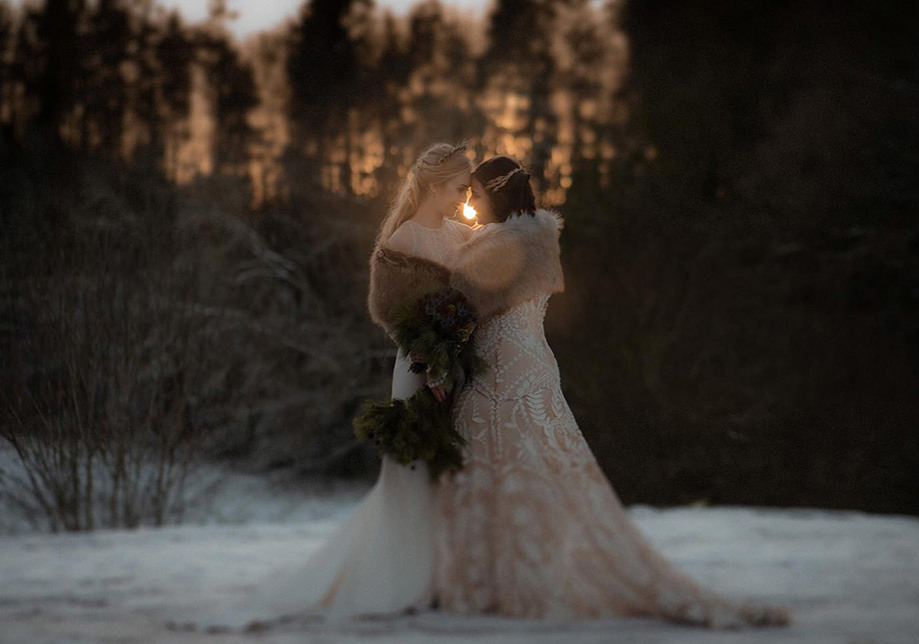 Two brides stand in snow wearing fur boleros embracing one another at sunset